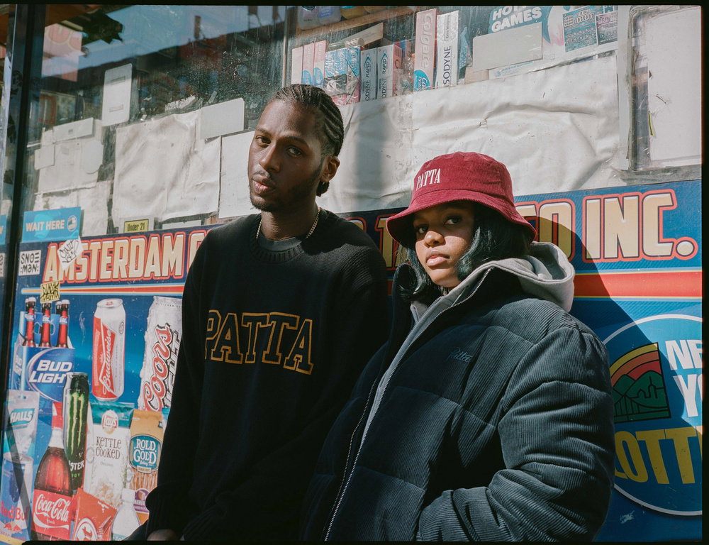 Two people pose in front of a colourful newsstand. One wears a black sweatshirt with “Patta” in gold lettering, while the other wears a red bucket hat and a dark puffer jacket.