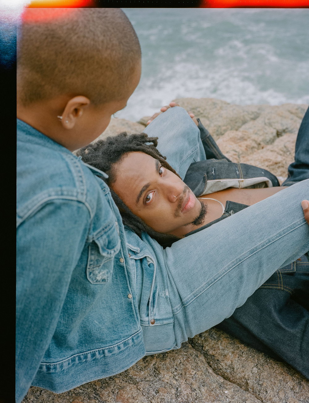 Two people lounge together on a rocky shoreline by the sea. One person rests their head on the other's lap, looking up at them. Both wear denim clothing.