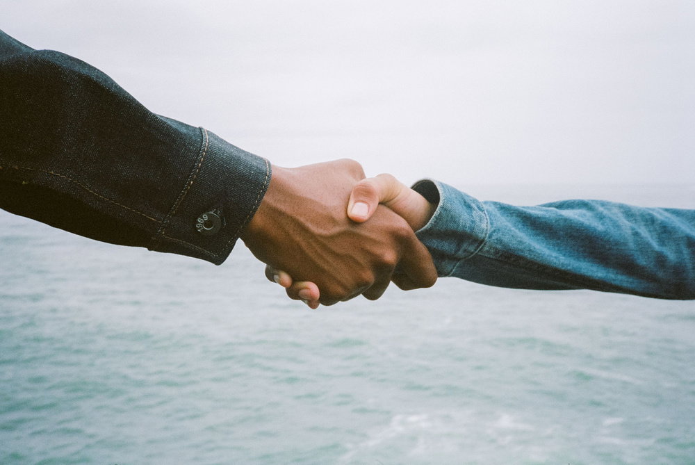 Two people shake hands against a backdrop of a pale sky and a choppy turquoise sea. Both wear denim jackets in different shades of blue.