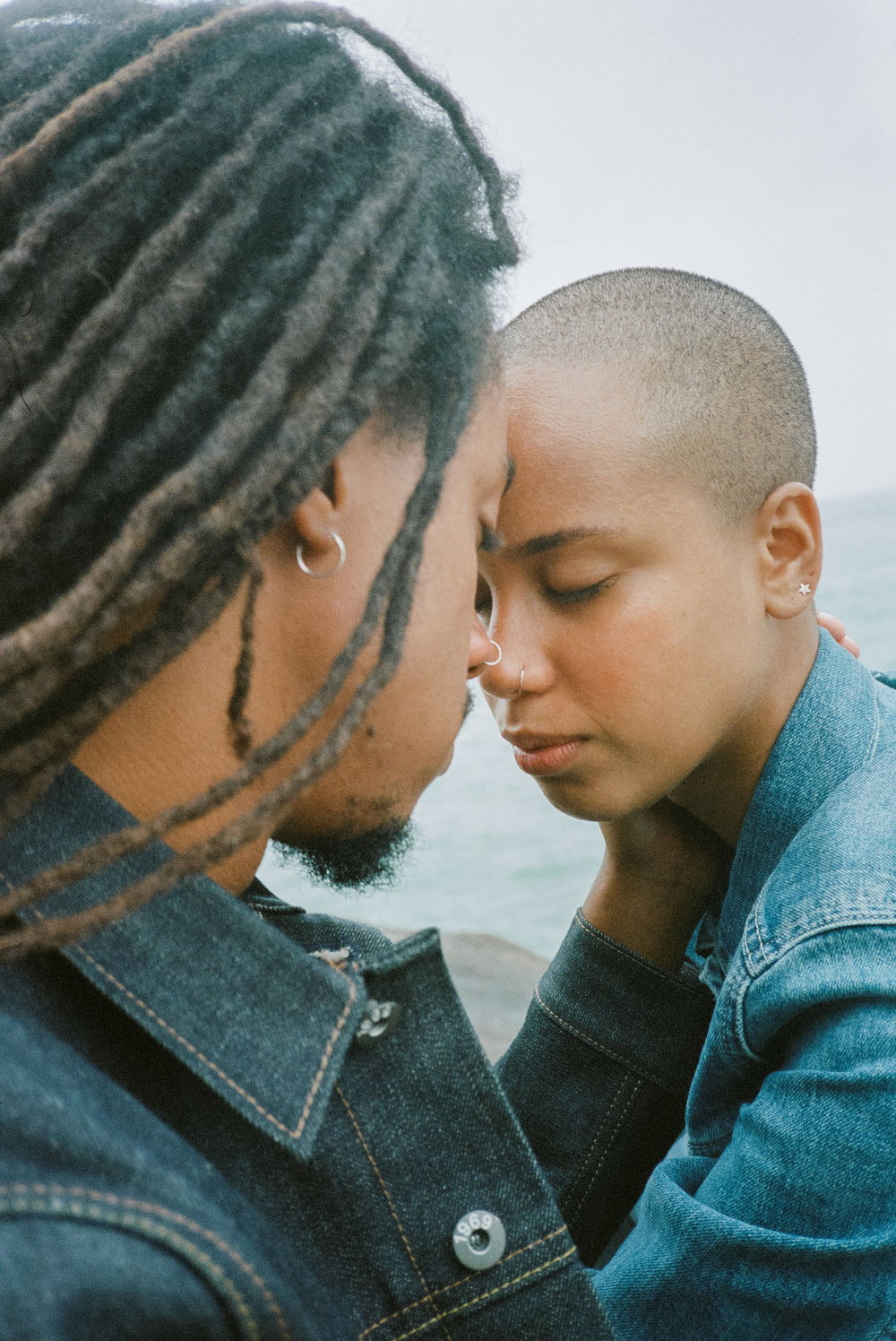 Two people lean in close with their foreheads touching and eyes closed. They both wear denim jackets against a soft, blurred outdoor backdrop.