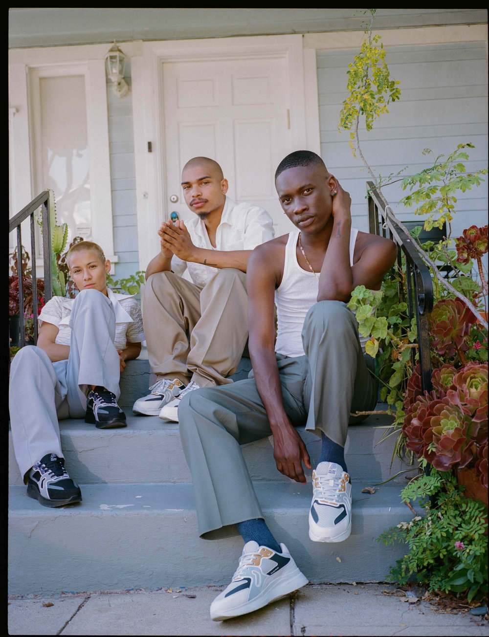 Three people sit on the steps of a white house, looking towards the camera. They wear neutral-toned clothing and chunky white and blue trainers.