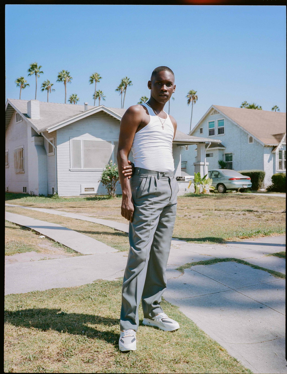 A person stands on a suburban lawn, looking directly at the camera. They wear a white ribbed tank top, grey trousers and white trainers. Behind them are light-coloured houses and tall palm trees under a clear blue sky.