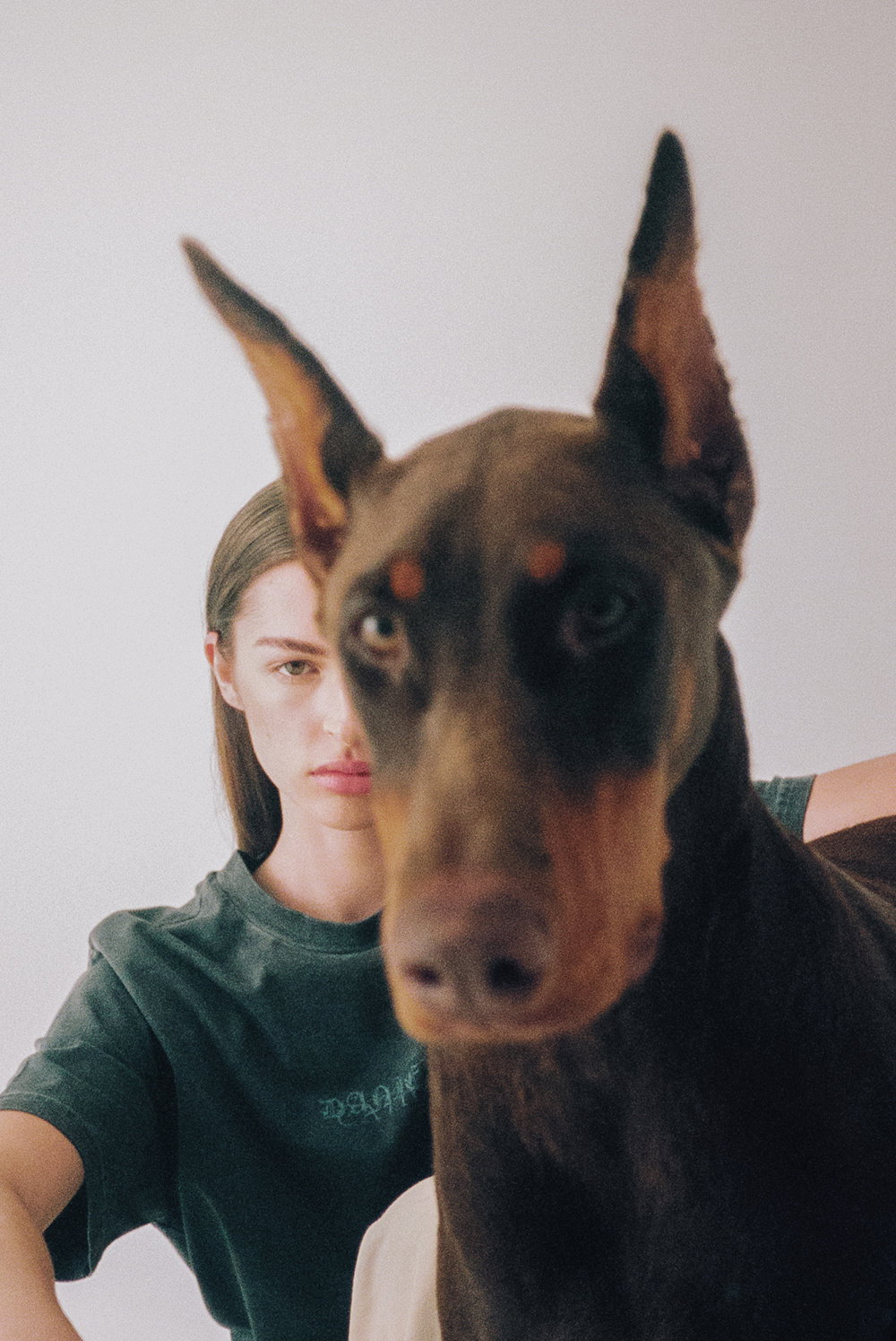 A Doberman Pinscher stands in the foreground, its face blurred and close to the lens. Behind the dog, a person in a dark green T-shirt looks directly at the camera with a neutral expression.