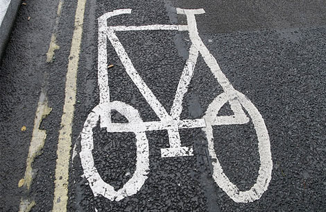 A white bicycle symbol is painted on a dark, textured asphalt road. Two yellow lines run parallel to the symbol along the left edge of the frame.