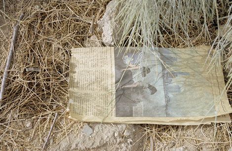 A weathered piece of newspaper lies on dry, sandy ground among dead grass and scrub. The page features a column of text and a faded photograph of a landscape.