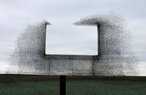 A sculpture of a large rectangular frame made from a dense, tangled mesh of wire. It stands on a single black post in an open field under a grey sky.