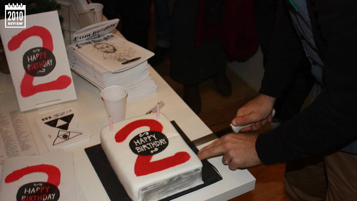 A person uses a knife to cut into a cake shaped like a stack of white papers. The cake is topped with a red number “2” and a black circle that reads “HAPPY BIRTHDAY”.