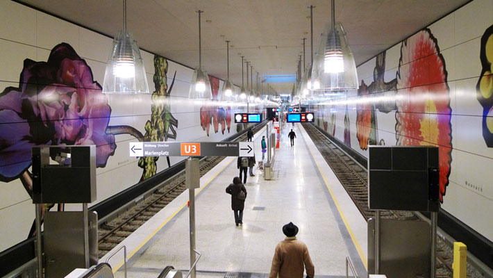 A wide shot of a clean, modern underground station platform. Large, colourful floral murals line the walls, with a person walking towards the tracks.