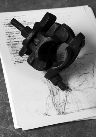 A heavy black metal clamp rests on a stack of sketches. The drawings beneath include handwritten notes and a gestural figure study.