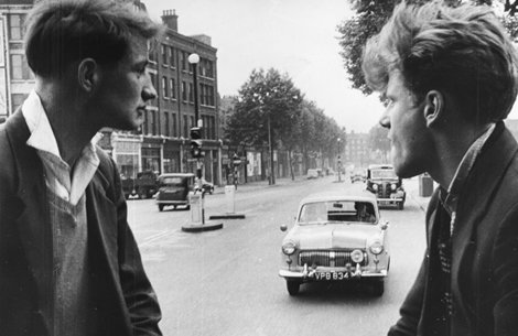Two people stand facing each other in profile on a city street. Behind them, vintage cars are parked and driving along a road lined with brick buildings and trees.