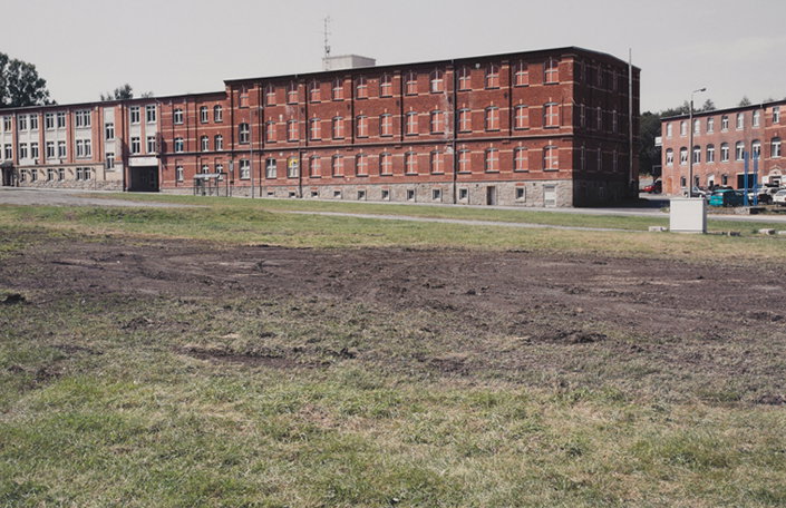 A long, three-storey red brick building stands behind a wide expanse of dirt and patchy grass. To the left, a connected section of the building is finished in a pale, weathered plaster.