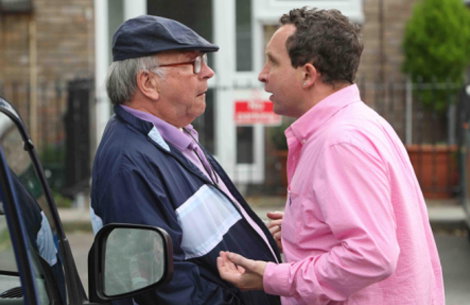 Two people stand close together in a heated conversation outdoors. One wears a navy blue jacket and a flat cap, while the other wears a bright pink shirt.