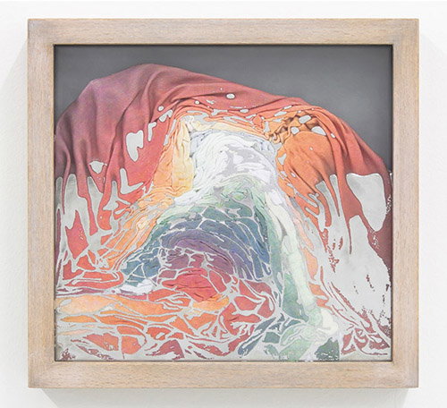 A framed piece of fabric art featuring a draped, mound-like shape in shades of red, orange and pale green. The material is layered and folded, creating a fluid, organic form against a dark grey background.
