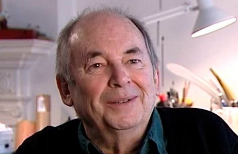 A person smiles warmly while looking slightly off-camera. They are in a bright room with a white desk lamp and studio equipment in the background.