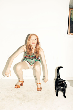 A person with long red hair crouches in a wide stance, looking directly at the camera. Beside them, a black cat stands with its tail curled upwards.