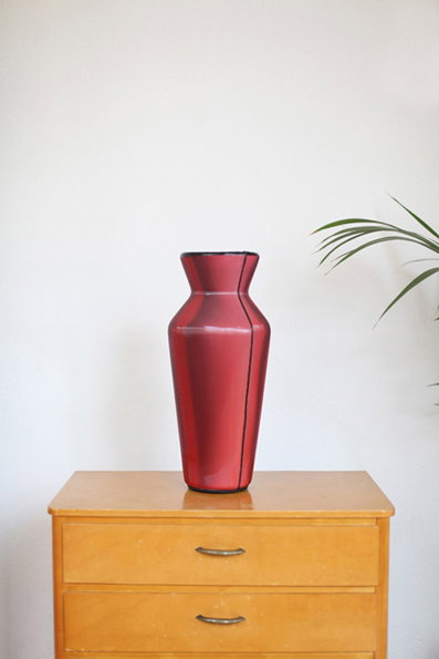 A tall, red glass vase with a tapered waist sits on a light wood chest of drawers. A few green palm leaves frame the right side of the image against a plain white wall.