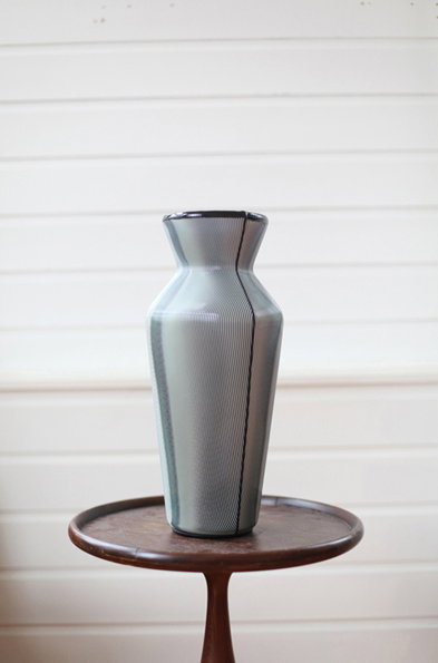 A slender, tapered vase with a fine vertical ribbed texture sits on a small wooden pedestal table. The vase is a muted blue-grey with a dark rim, set against a backdrop of white horizontal wall panels.