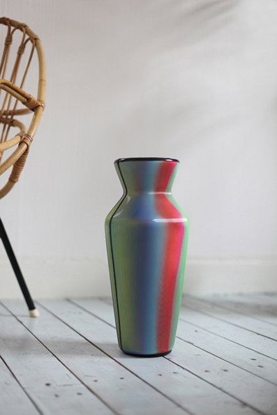 A iridescent vase with a gradient of blue, green and red hues stands on a light wood floor. To the left, the curved frame of a rattan chair is partially visible.