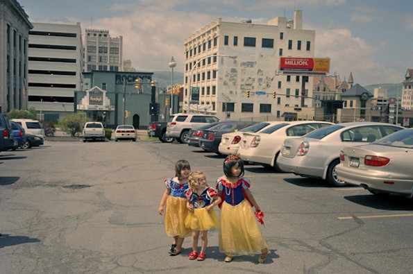 Three children dressed as Belle from Beauty and the Beast stand in the middle of a cracked city road. Behind them, a row of parked cars lines the street against a backdrop of urban buildings.