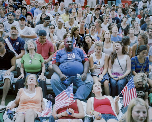 A large crowd of people sit in bleachers, many wearing blue and red clothing. In the centre, a person with blue and red face paint wears a blue T-shirt with a “ny” logo.