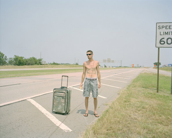 A shirtless person wearing sunglasses and cargo shorts stands on the shoulder of a wide, empty highway. A wheeled suitcase stands beside them next to a speed limit sign that reads “SPEED LIMIT 60”.