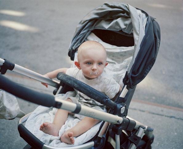 A child sits in a grey and black pram, looking off to the side. They have a small smudge of white food on their chin.