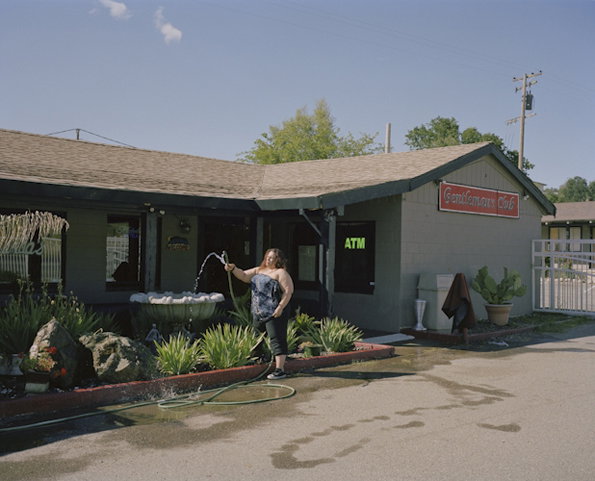 A person stands outside a grey building, holding a hose and spraying water into a stone basin. A red sign on the building reads “Gentlemans Club”, with a green “ATM” sign nearby.