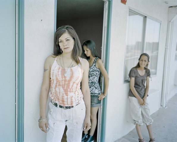 Three people stand in and around a doorway of a plain white building. One person stands in the foreground looking at the camera, while two others linger behind them.