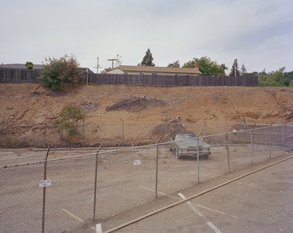 A green car is parked in a fenced-off concrete lot. Behind it, a dirt embankment leads up to a wooden fence and a single-storey house.