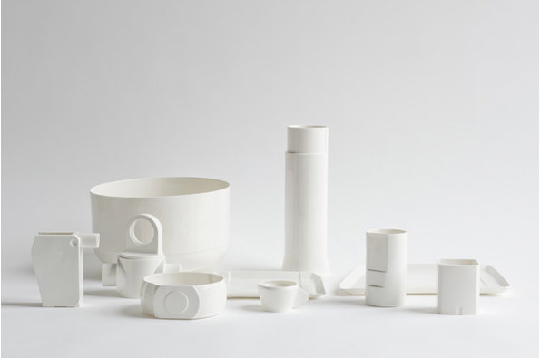 A collection of white ceramic vessels and geometric forms arranged against a plain light grey background. The pieces vary in height and shape, including a large wide bowl, a tall cylinder, and several smaller cups and trays.