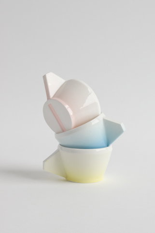 Three ceramic cups in pastel pink, blue and yellow are stacked precariously against a plain white background. The cups are tilted at different angles, creating a balanced but unstable tower.