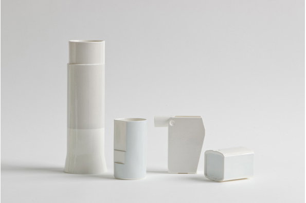 Four white ceramic vessels of varying heights and geometric shapes stand in a row against a plain grey background. The tallest piece is a slender cylinder, while the others are shorter and more angular.