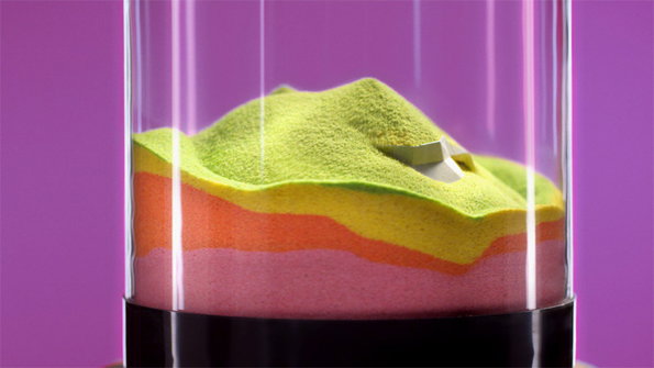 Layers of brightly coloured sand in pink, orange, yellow and green are contained within a clear glass cylinder. A small white object is partially buried in the top green layer.