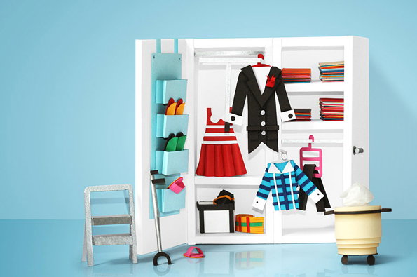 A miniature white wardrobe is open to reveal a collection of tiny clothes and accessories. A black tuxedo, a red and white striped dress, and a blue checked shirt hang inside alongside folded fabrics and shoes.