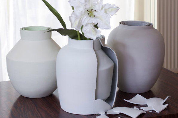 Three matte white ceramic vases sit on a dark wooden table. The central vase is cracked open, with a white flower emerging from the top and shards of ceramic scattered around its base.