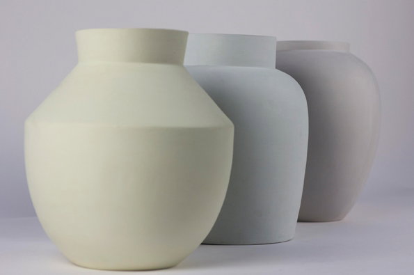 Three matte ceramic vases in cream, light grey and pale mauve stand in a row. They feature soft, rounded shapes and flat rims against a plain white background.