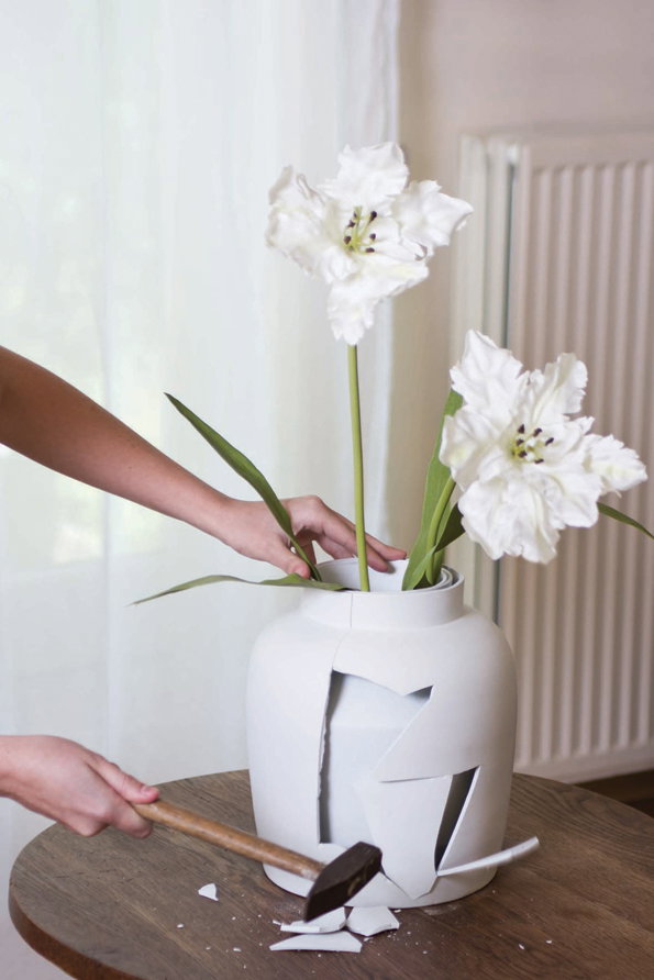 A person uses a wooden mallet to smash a white ceramic vase on a wooden table. Two large white flowers remain standing in the vase as it breaks into shards.