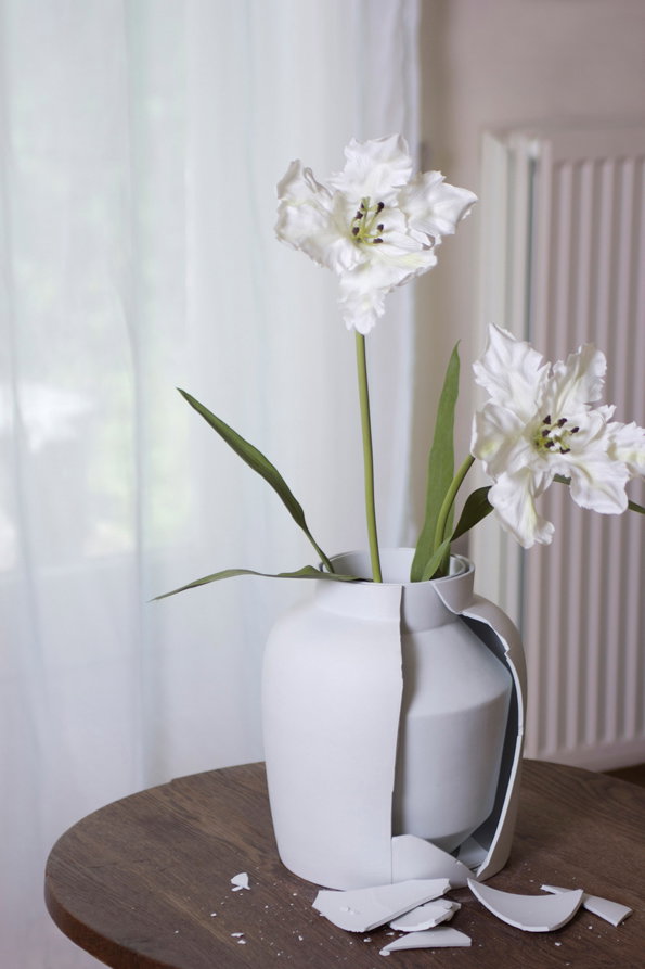 Two white lilies stand in a matte white ceramic vase. The vase is cracked and broken, with several shards resting on the wooden table below.