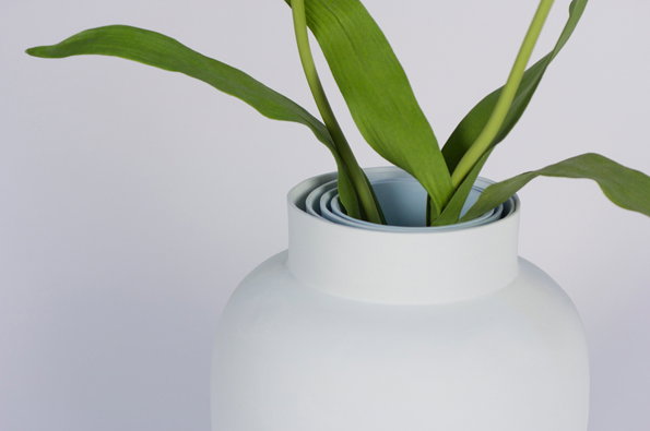 A white ceramic vase holds several long, green leaves that arch outwards. The vase has a tiered, concentric rim around the opening.