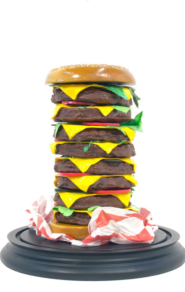 A sculpture of an impossibly tall burger with seven patties, melted yellow cheese, and slices of tomato. It sits on a black circular plinth, wrapped at the base in red and white checkered paper.