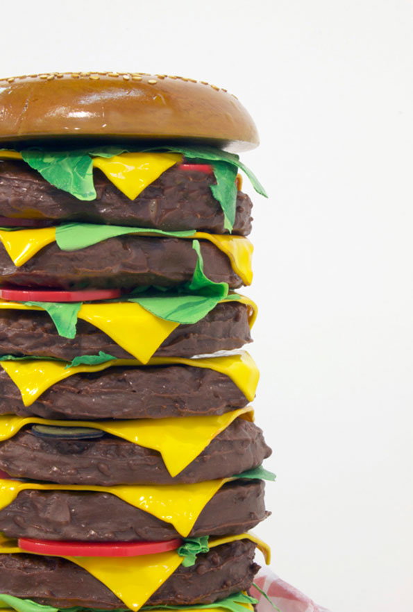 A sculpture of a towering burger with six brown patties, yellow cheese slices and green lettuce. The oversized stack is topped with a glossy, sesame-seeded bun against a plain white background.