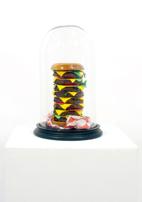 A sculpture of an oversized burger with multiple patties, cheese slices and lettuce. It sits on crumpled red paper under a glass cloche on a white plinth.