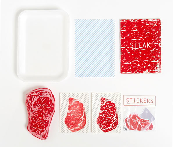 A flat lay of a stationery set inspired by raw meat. The items include a white tray, a blue striped card, a red textured card reading “STEAK”, and several stickers of marbled red meat.
