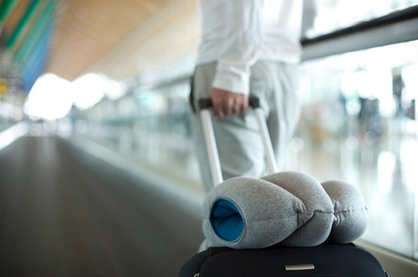 A person pulls a suitcase along a moving walkway in an airport. A grey travel pillow rests on top of the black luggage.