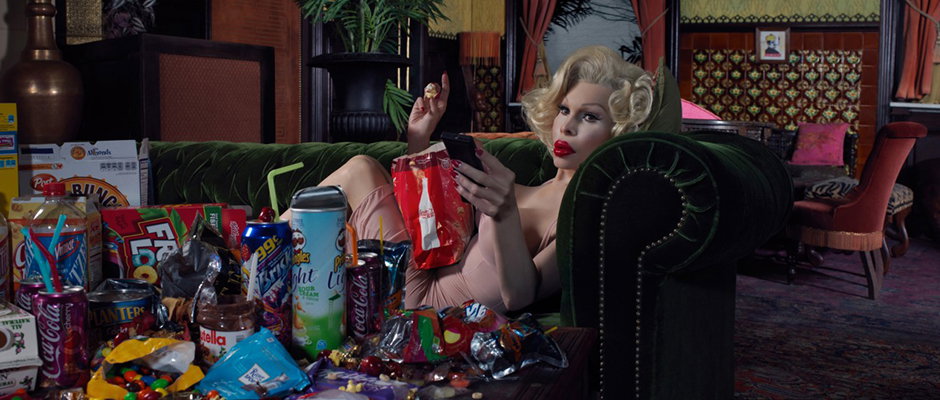 A person styled as Marilyn Monroe reclines on a green velvet sofa, looking at a smartphone. They are surrounded by an abundance of open snack bags, drink cans and fast food containers.