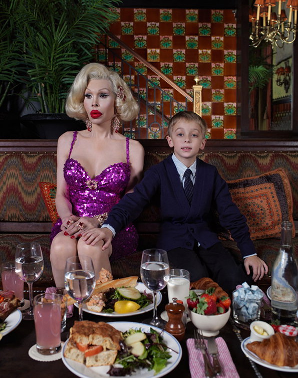 A person in drag wearing a sparkling purple dress and blonde wig sits beside a child in a navy suit. They sit on a patterned sofa behind a table laden with croissants, salads and drinks.