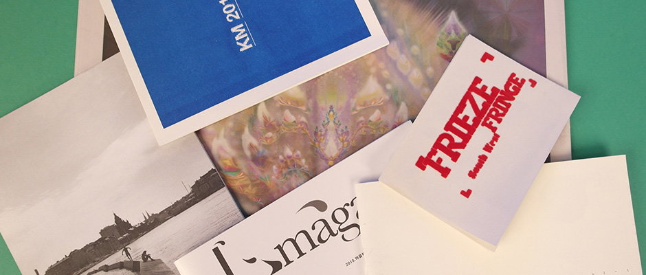 A collection of magazines and pamphlets are scattered across a green surface. One white booklet features the word “Frieze” in bold red lettering, while another shows a black and white landscape.