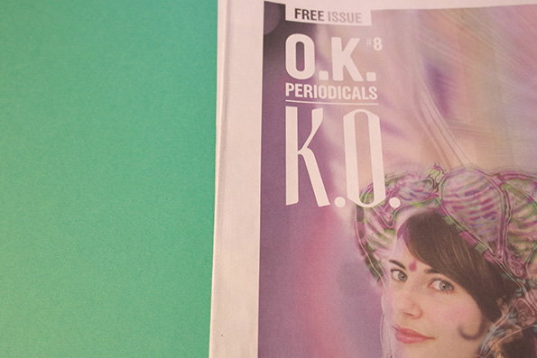 The cover of a magazine titled “O.K. Periodicals” features a person wearing a large, iridescent, shell-like headpiece. The text “Free Issue” and “K.O.” appear in bold white lettering over a purple and pink gradient background.