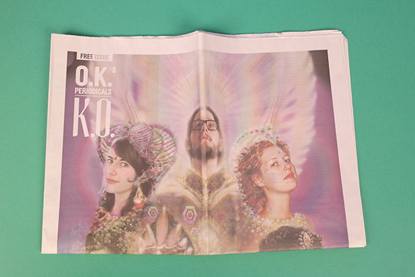 The cover of a magazine titled “O.K. Periodicals” features three people in ornate, shimmering costumes. They are set against a soft, glowing purple and pink background.