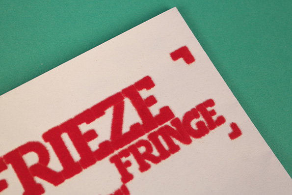 A close-up of a white card on a green background. Bold red text reads “FRIEZE FRINGE”.